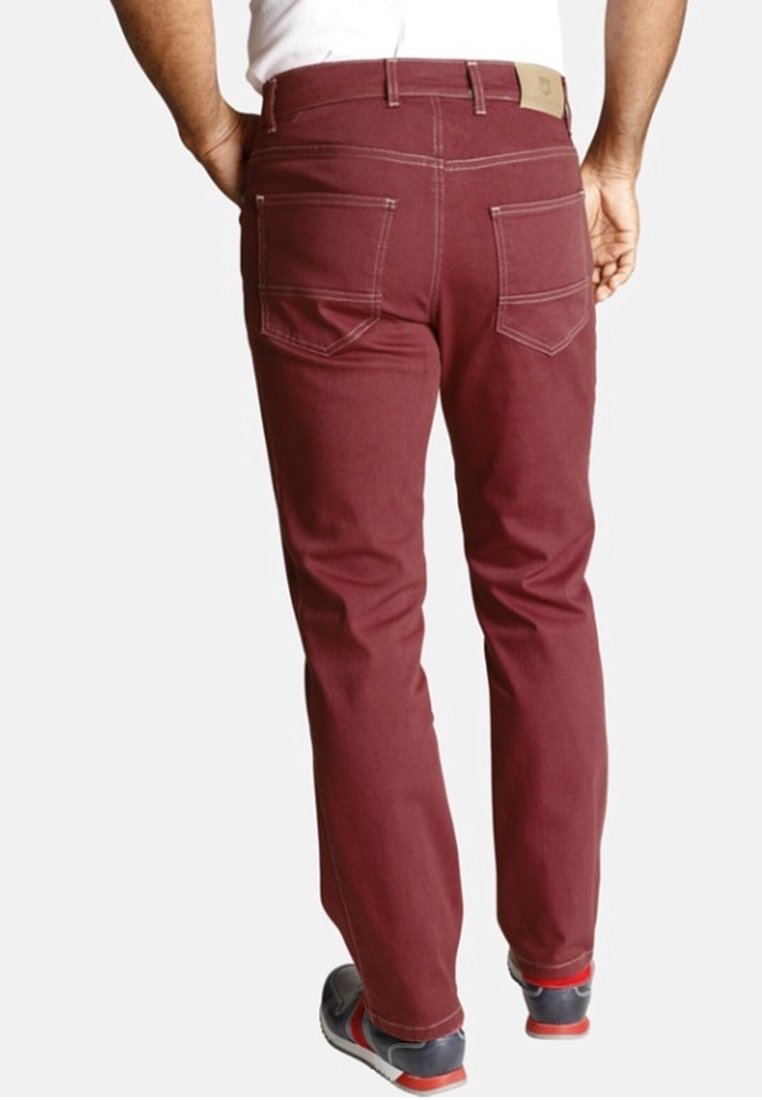 Homme portant un pantalon slim marron avec des poches arrière visibles, une chemise blanche et des baskets foncées avec des accents rouges, debout devant un fond blanc.