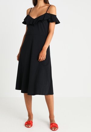 Kvinde iført en sort off-shoulder midi-kjole med flæser og røde flade sandaler, stående mod en ensfarvet baggrund.