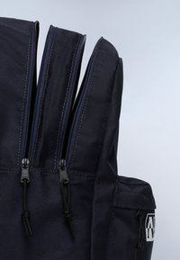 Zaino blu navy con molteplici scomparti con zip, tessuto resistente e accessori neri. Presenta un cordino regolabile per una chiusura sicura.