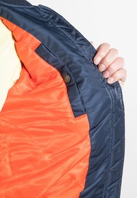 Granatowa pikowana kurtka z jasnopomarańczową podszewką, ukazująca gładką teksturę oraz zatrzask na wewnętrznym szwie.