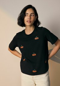 T-shirt en coton noir avec des motifs de citrouilles brodées en orange de différentes tailles, col rond et manches courtes.