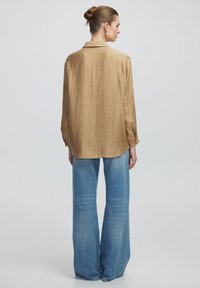 Bluse mit bronzefarbenem Textur und Kragen, kombiniert mit hellblauen, hochgeschnittenen Schlagjeans. Der Stoff hat ein zartes, zerknittertes Finish.