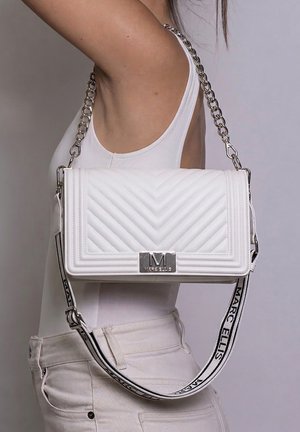 Witte gewatteerde schoudertas met zilveren kettingriem en logoplaatje, gedragen door een persoon in witte top en broek.