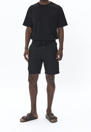 Hombre con camiseta negra, pantalones cortos con cordón negros y sandalias marrones, de pie contra un fondo blanco.