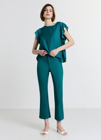 Blouse turquoise à volants avec une texture lisse, associée à un pantalon turquoise ajusté. Des talons blancs ouverts complètent la tenue.