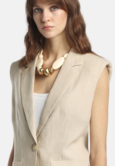 Tânără cu păr ondulat castaniu, purtând un sacou bej fără mâneci, bluză albă și un colier mare crem cu detalii aurii.