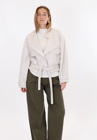 Femme aux cheveux longs portant une veste surdimensionnée beige clair ceinturée, un haut blanc et un pantalon ample vert olive, debout devant un fond blanc uni.