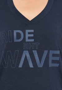 Camiseta navy de cuello en V con letras texturizadas que dicen "CABALGAR LA OLA" en azul claro con diferentes tamaños y estilos de fuente. Tejido suave, corte relajado.