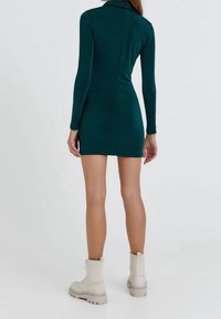 Robe verte à manches longues avec une silhouette ajustée et un col, vue de dos. Associée à des bottes de combat beiges. Texture lisse.