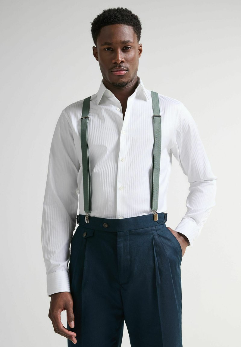 Camisa de dress blanca a rayas acompañada de pantalones azul oscuro, con tirantes verdes y clips metálicos. Corte slim, textura suave.
