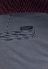 Pantalones de tela gris con una cintura de canalé color burdeos oscuro, un bolsillo lateral y una etiqueta negra con la marca cosida en la cintura.