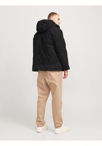 Zwarte capuchonjack met een gladde textuur, tweekleurig ontwerp en een verlengd achterpand, gecombineerd met beige broeken en witte sneakers.