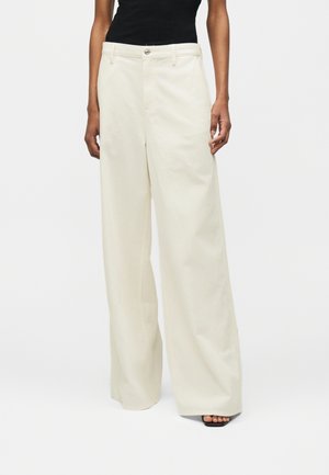 Femeie purtând pantaloni crem cu talie înaltă și picioare largi, cu închidere cu nasture și fermoar, asortați cu sandale negre cu toc și degetele deschise.