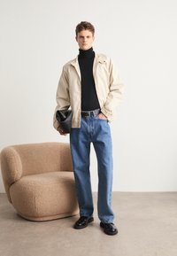 Beige Jacke über einem schwarzen Rollkragenpullover, kombiniert mit blauen Jeans, schwarzen Schuhen und einer schwarzen Clutch. Texturierter brauner Stuhl im Hintergrund.