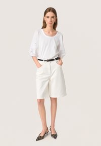 Hvid bluse med puffede ærmer og øjelet detaljer, parret med knæ-lange hvide shorts. Accentueret med et sort bælte og zebra-mønstrede hæle.