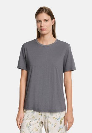 Kobieta z prostymi włosami, ubrana w jednolitą szarą koszulkę z krótkim rękawem oraz jasne spodnie z abstrakcyjnymi pastelowymi wzorami, stojąca przodem.