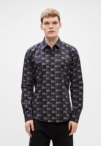 Μακρυμάνικη μπλούζα από μαύρο ύφασμα με επαναλαμβανόμενο μοτίβο γραφικών «HUG» σε διάφορες αποχρώσεις του γκρι και του ροζ, με κλασικό γιακά και κουμπί.