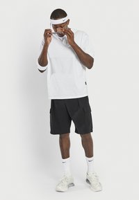 T-shirt blanc à manches courtes, shorts cargo noirs, bandeau blanc, lunettes de soleil de sport et baskets blanches avec des accents gris. Tenue athlétique décontractée.