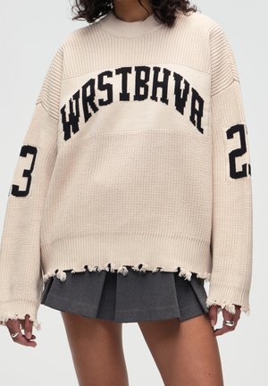 Suéter beige de gran tamaño con texto negro "WRSTBHVR", puños y dobladillo desgastados, combinado con falda plisada gris oscuro.