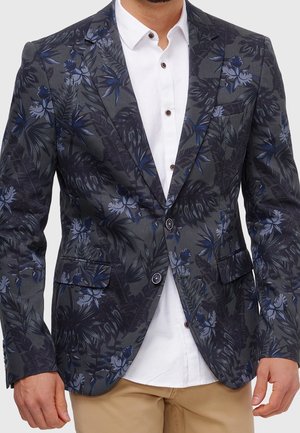 Mann trägt dunkelgrauen Blazer mit blauem Blumenmuster, weißes Hemd mit Knöpfen und beige Hose vor weißem Hintergrund.
