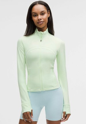 Vrouw met lang donker haar die een lichtgroen sportjack met rits en lichtblauwe nauwsluitende shorts draagt, staand tegen een witte achtergrond.