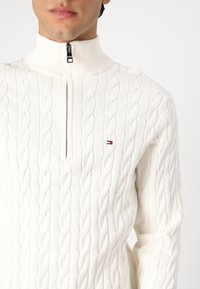 Witte gebreide trui met kabelpatroon, hoge kraag en halve ritssluiting. Bevat een klein logo in rood, wit en blauw op de borst.