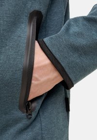 Teal fleecejack met een zwart ritszakdetail. Gelaagd materiaal en slanke manchetten zorgen voor extra functionaliteit en pasvorm.