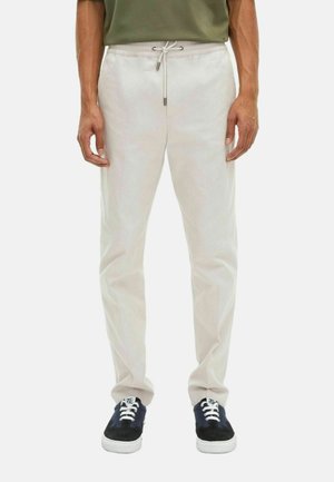 Man in lichtbeige joggingsbroek met een trekkoord en zwart-blauwe sneakers, staand tegen een witte achtergrond.