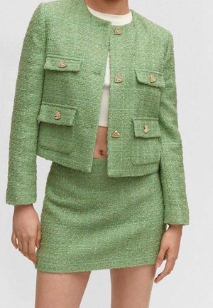 Femme portant une veste courte en tweed vert avec des boutons dorés en forme de cœur et une mini-jupe taille haute assortie, montrant le ventre et les mains.