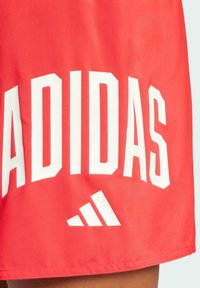 Červené atletické šortky vyrobené z ľahkej tkaniny, so silným bielym textom a logom "ADIDAS" v geometrickom serifovom písmene. Hladká textúra.