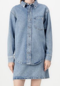 Light blue denim shirt with long sleeves, button closure, chest pocket, and curved hem, paired with a matching denim skirt.