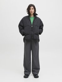 Veste noire matelassée avec col haut, deux poches avant, associée à un haut vert et un pantalon gris ample. Le modèle porte des chaussures noires épaisses.
