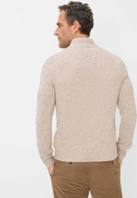 Strickpullover in hellbeige mit strukturiertem Muster, hohem Kragen und gerippten Bündchen. Kombiniert mit braunen Cordhosen, die einen lässigen Look zeigen.
