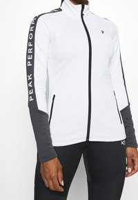 Witte zip-up jas met hoge kraag, grijze accenten op de mouwen, voorzien van de tekst "PEAK PERFORMANCE" langs de arm en ritszakken.