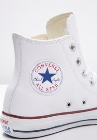Adidași albi de tip high-top, din piele netedă, cu un logo rotund albastru cu o stea, ochiuri argintii și detalii roșii pe talpă.