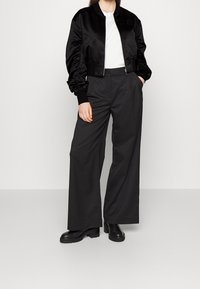 Zwarte satijnen bomberjack met gathered mouwen, gecombineerd met brede zwarte broeken. Model draagt een wit t-shirt eronder. Stevige zwarte laarzen.