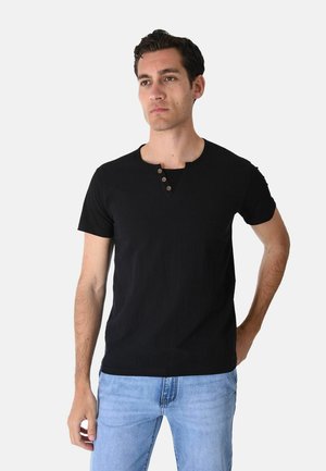 Uomo che indossa una maglia henley nera a maniche corte con tre bottoni, jeans azzurri, in piedi con una mano in tasca su uno sfondo semplice.