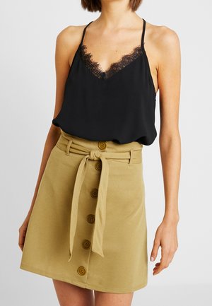 Femme portant un caraco noir orné de dentelle, rentré dans une jupe beige à boutons sur le devant, avec une ceinture nouée à la taille.