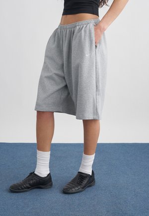 Osoba nosząca szare, sięgające do kolan spodenki Nike, czarne trampki, białe skarpetki i czarny krótki top, stojąca na niebieskim dywanie.