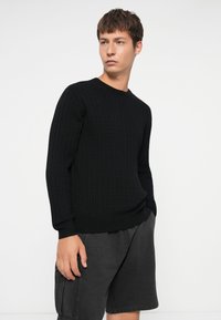 Sort strikket sweater med ribbet tekstur, rund halsudskæring og lange ærmer, sat sammen med mørke shorts. Enkel, klassisk design.