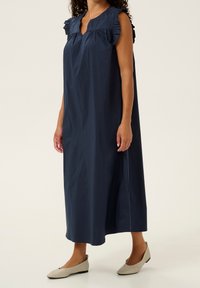 Mörkblå klänning med volangdetaljer på axlarna, knälång, tillverkad av lätt tyg. Kombinerad med beige platta skor.