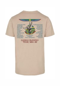 Beige bomull t-skjorte med et grafisk motiv av en ankh og et hundehode, med navnet på landene trykket i blått, og "WORLD SLAVERY TOUR 1984-85" i rødt.