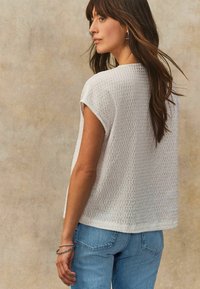 Bílý, krátký rukáv, texturovaný top s volným střihem, v kombinaci se světlými modrými džíny, zdůrazňující uvolněnou siluetu a decentní detaily.