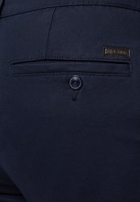 Granatowe spodnie o gładkiej fakturze, z tylną kieszenią zabezpieczoną guzikiem oraz markową skórzaną metką na pasku.