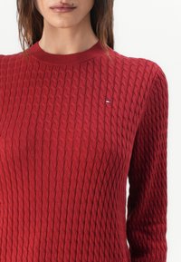 Jersey de punto rojo con un diseño de trenzas texturizado, que presenta un cuello redondo y un pequeño logo de marca en el lado izquierdo del pecho.