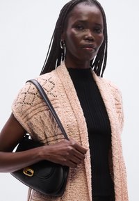 Μαύρη δερμάτινη τσάντα χειρός με χρυσή αγκράφα. Το μοντέλο φοράει ένα ροζ πλεκτό ζακέτα με υφή πάνω από ένα ριμπ μαύρο τοπ, παρουσιάζεται με φόντο ουδέτερου χρώματος.