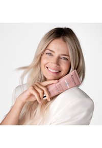 Smilende kvinde med langt blondt hår holder en lyserød Kérastase Chroma Absolu hårplejetube op til sit ansigt, iført en hvid blazer.