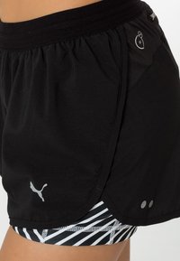 Svarta sportshorts med bred midja, kontrasterande inre mesh-lager med ett vitt randmönster och silverfärgade logotypaccenter.