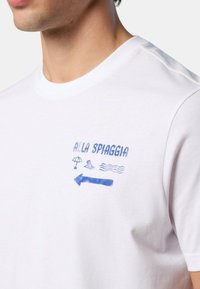 T-shirt in cotone bianco con grafiche blu raffiguranti un ombrello, un uccello, onde e una freccia con il testo "ALLA SPIAGGIA." Maniche corte, collo rotondo.