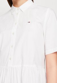 White short-sleeve shirt dress with a button-up front, gathered waist, and a small logo patch featuring red, white, and blue colors.